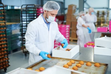 Confectionery factory worker in white coat collecting freshly baked pastry from tray and putting it into paper box.